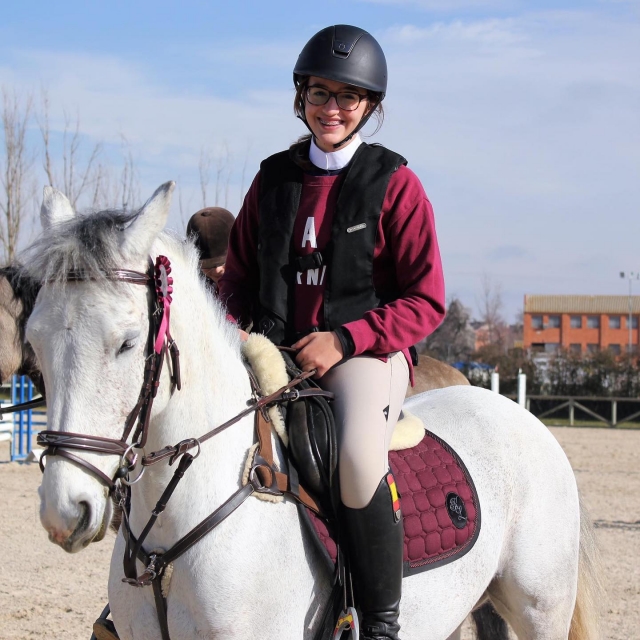  A smiling girl, with her respective protective helmet, on the back of a white horse 